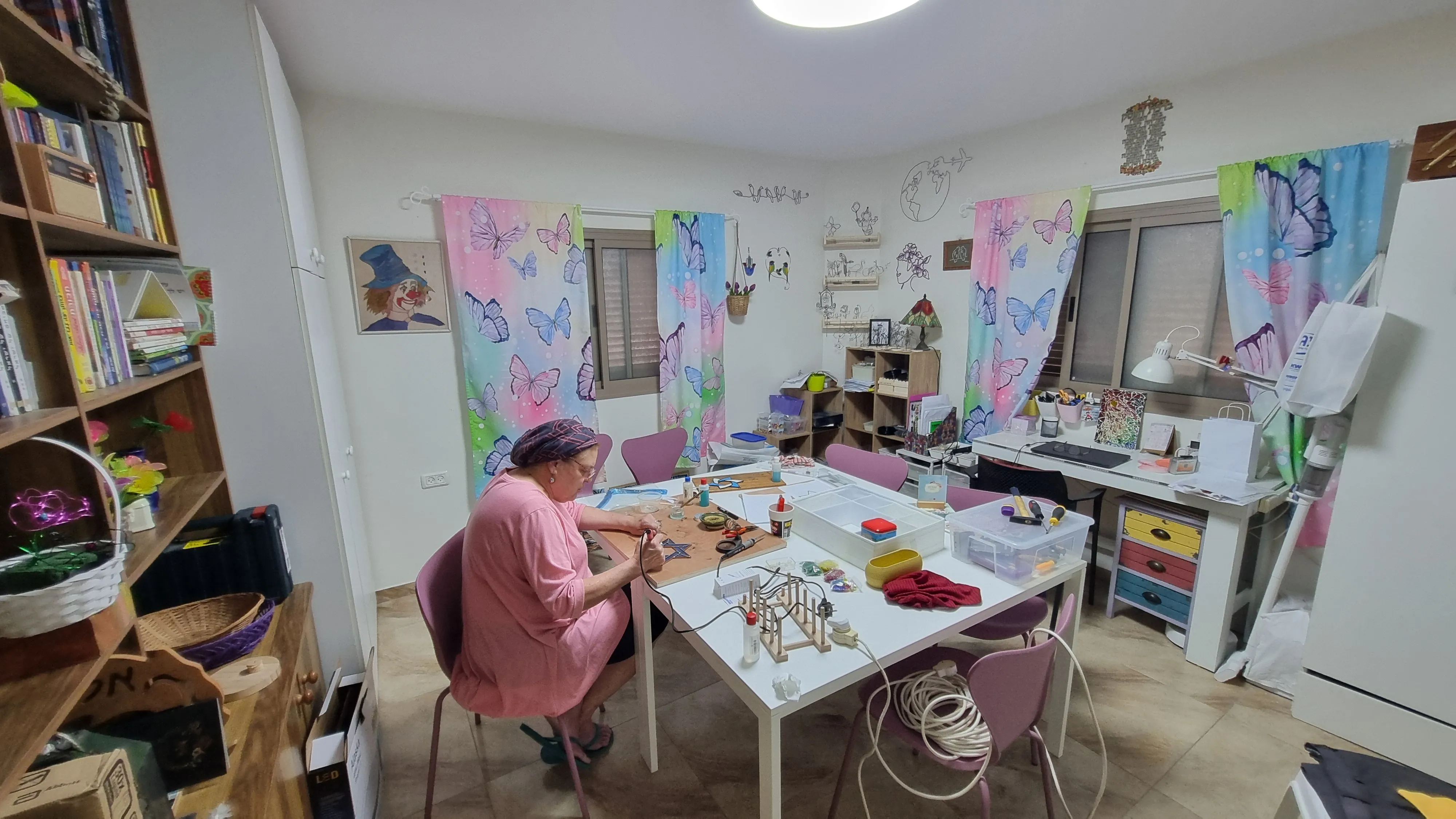 תמונה רחבה של הסטודיו ובו שרה יושבת מול השולחן ומלחימה זכוכית
